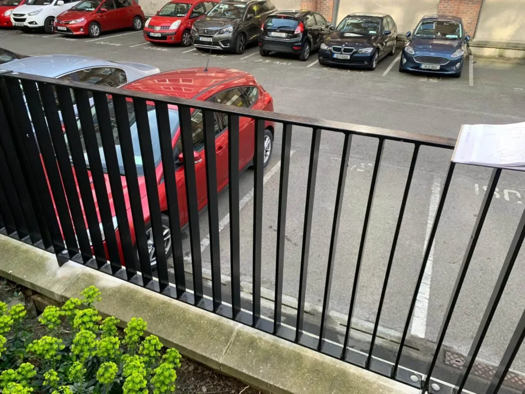 Cars parked in a lot seen through a black metal fence with green plants nearby.